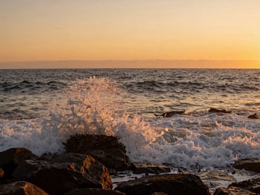 Wide coastal shot of Viña del Mar at golden hour, waves breaking against dark rock in the foreground, warm orange light on the water, low horizon, no people