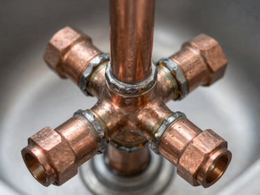 Overhead close-up of copper plumbing connections under a sink, soldered joints and pipe fittings in sharp detail, diffused natural daylight, no staging