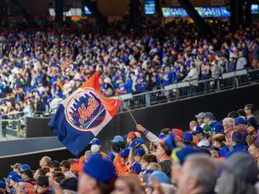a stadium full of fans waving a large orange and blue mets flag at 2024 nlcs game 5