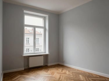 Interior corner of a renovated Klaipėda apartment, pale grey walls, parquet floor, large window with diffused daylight revealing a street below, minimal furniture, shot from entry angle