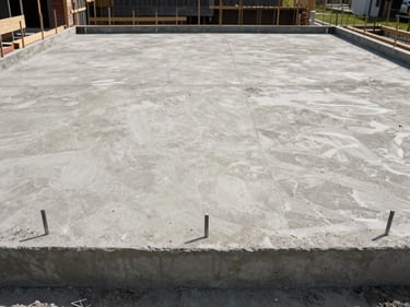 Straight-on daylight shot of a finished concrete residential foundation and ground-floor slab — formwork removed, level surface visible, construction stakes still in place at perimeter