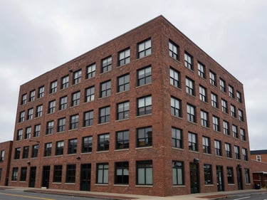 Wide environmental shot of a mid-rise commercial building exterior, direct overcast daylight revealing brick coursework and window rhythm, street-level framing showing full facade scale, neutral sky