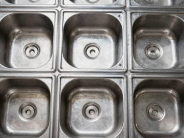 Tight overhead shot of stainless steel sink blanks arranged in a factory grid, studio strobe, even industrial lighting, showing gauge thickness and weld seams, no warmth or atmosphere