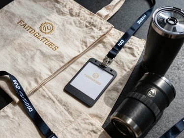 Tight overhead shot of custom branded merchandise — embroidered tote bag, lanyard, and tumbler arranged on a dark surface under north-facing daylight, close crop revealing stitch texture and foil detail on each piece