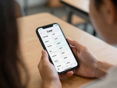 Learner reading from a phone with English vocabulary flashcards on screen, seated at a wooden table under diffused classroom light