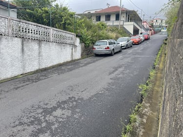 Cars parked along a narrow uphill residential street with white stone walls and houses.