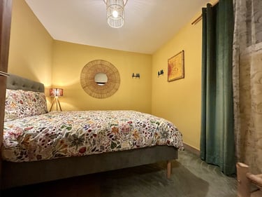 Cozy bedroom with yellow walls, floral bedding, a wicker mirror, and soft ambient lighting.