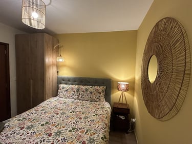 Cozy bedroom with floral bedding, woven wall mirror, and warm ambient lighting from bedside lamps.