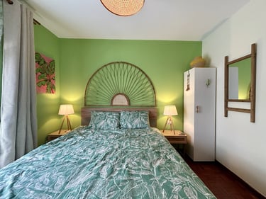 Tropical bedroom interior featuring green walls, a rattan headboard, and leafy print bedding with warm bedside lamps.