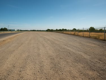 Light-industrial zoned land plot with gravel surface, perimeter fencing partially visible, clear afternoon sky, wide shot showing lot depth and access road at left