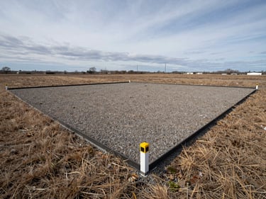 Flat residential land parcel in daylight, survey boundary markers at corners, dry grass and gravel surface, clear overcast sky, wide environmental framing