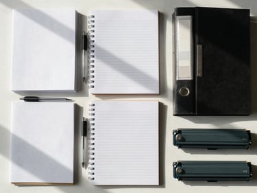 Overhead flat-lay of office supplies on a clean desk — reams of paper, pens, binders and toner cartridges arranged in a precise grid under north-facing daylight, functional and orderly