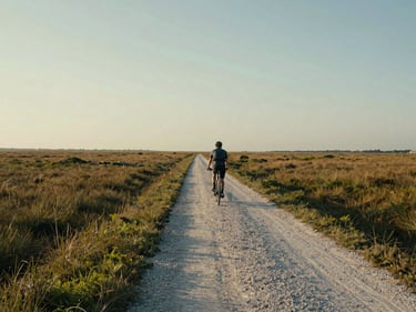 Wide environmental shot of a lone cyclist on a flat gravel path cutting through a salt marsh at golden hour, the path running to the distant horizon, vast sky above, muted greens and amber grasses either side, no posing, documentary framing