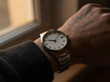 Close-up of a minimalist steel watch on a man's wrist, textured dark knit fabric sleeve, warm side lighting, shallow depth of field.