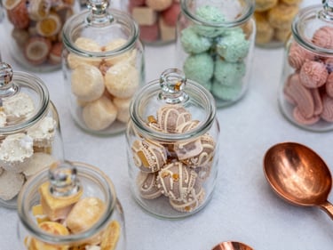 Extreme close-up overhead of a styled candy table surface: glass apothecary jars filled with pastel confections, copper scoops resting on white linen, bright natural daylight, shallow depth of field revealing jar texture and ribbon detail
