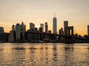 The iconic skyline of New York City from Brooklyn Bridge Park during sunset, warm golden light reflecting on the water, editorial travel photography, 35mm lens