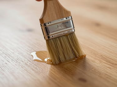 Extreme close-up of a brush applying water-based lacquer to a smooth ash floor surface, tungsten light catching the wet film and revealing grain texture beneath the first coat
