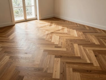 Wide environmental shot of freshly installed herringbone teak floor in an empty room, sidelight entering from a tall window at left, subfloor expansion gaps visible at the perimeter, no furniture