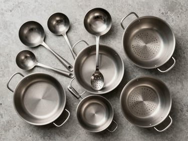 Overhead flat-lay of stacked kitchen equipment — ladles, pans, and colanders arranged on a neutral concrete surface, clean studio light, clear texture and material detail