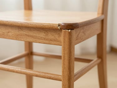 Close-up of a broken wooden chair leg, clearly damaged and unstable, in soft indoor lighting.