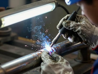 Close-up of a pipe weld joint being inspected under bright overhead fluorescent light, welder's gloved hands holding inspection tool against the joint, sharp documentary framing