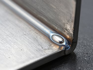 Extreme close-up of a TIG weld bead on a stainless steel sheet corner, weld dressed and polished to blend, cold white studio lighting at a low angle that reveals surface flatness and bead geometry