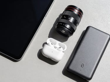 Flat-lay overhead shot of consumer electronics — a tablet, wireless earbuds case, and power bank arranged on a neutral gray surface, bright north-facing daylight, no branded packaging, product condition clear