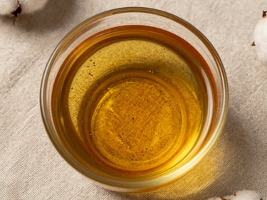 Extreme close-up overhead flat-lay of raw pressed cottonseed oil in a clear glass vessel on rough linen cloth, showing amber liquid clarity and oil surface texture, studio strobe from above, evidential framing