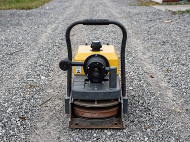 Operator-angle shot of a plate compactor on a gravel surface, viewed from behind-above, compactor plate and handle visible, diffused daylight from an open yard, no people