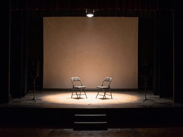 Small bare stage with two folding chairs and a single work light, viewed from the wings, deep shadows framing the lit performance area