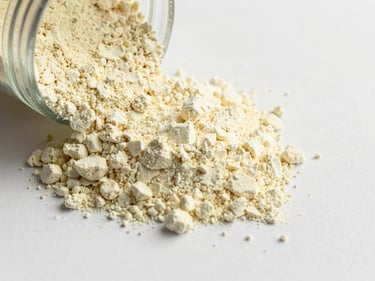 Close-up of pale yellow lyophilized brightening powder poured onto a matte white ceramic surface, fine granular detail visible, cool even daylight-balanced studio lighting