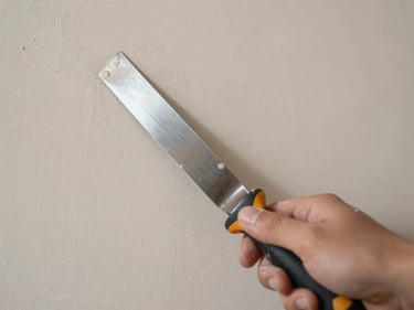 Overhead close-up of fresh plaster being applied to a drywall panel, wide joint knife in frame, natural studio daylight, neutral warm tones, no people visible
