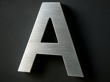 Close-up of a brushed aluminium 3D sign letter mounted on a dark wall, cool daylight raking across the raised surface revealing depth and edge detail