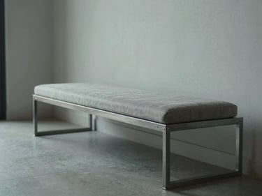 Wide panoramic of a steel bench with upholstered seat in a minimal residential entryway, cool north daylight, steel frame with visible joinery, one hand resting on the frame, tactile detail visible, generous negative space
