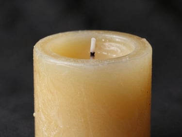 Side-lit macro of a thick beeswax taper candle against a dark linen backdrop, wax surface showing hand-poured texture and subtle drips, warm golden studio light catching the ridged surface, ivory and deep shadow tones