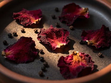 Extreme close-up of dried rose petals and black pepper corns scattered across dark terracotta ceramic, golden-hour side light raking across the surface revealing shadow and texture, burgundy and rust tones against near-black ground