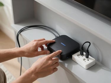 Overhead view of hands connecting a smart home hub to a power socket, device cables neatly arranged on a light grey shelf, natural daylight from the left