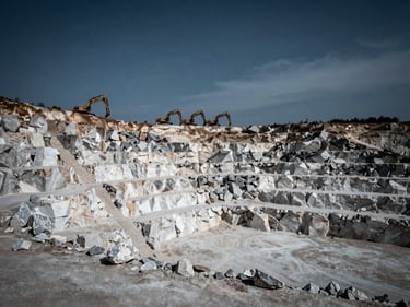 Wide shot of a massive limestone quarry, showing layers of white and grey rock, with heavy excavators in the background under an overcast sky