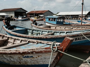 Close-up of a Balikpapan harbor at midday, wooden fishing boats moored in a row, textured rope and weathered hull in foreground, port warehouses visible behind, natural daylight, documentary framing