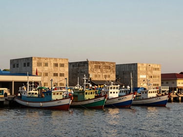 Wide shot of Balikpapan waterfront at late afternoon, fishing boats moored in the foreground, warm concrete port buildings behind, natural golden hour light, no filters, documentary framing
