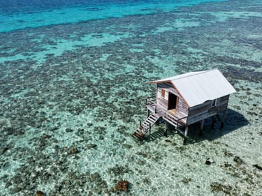 Close aerial view of San Andrés sea of seven colors, shallow reef visible beneath crystalline water, wooden stilted cabin to the right, bright midday Caribbean light
