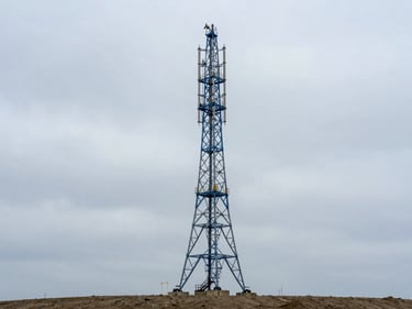Mid-range environmental shot of a completed three-legged steel cell tower rising against an overcast sky, royal blue safety equipment visible on the lattice legs, wide site context showing cleared ground below, daylight