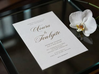 Overhead composition of an elegant event invitation displayed on a dark glass surface — intricate typographic layout glowing softly on screen, a single white orchid bloom placed at the frame's edge, moody studio lighting with turquoise reflection