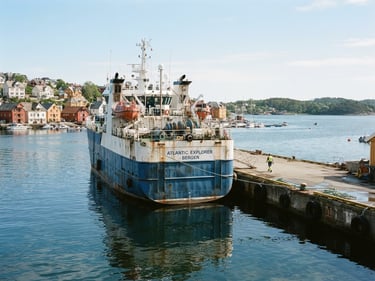 Medium environmental shot of a commercial transport vessel docked at a quiet harbor, bright natural daylight reflecting off the calm water, steel hull details visible, 35mm crisp focus.