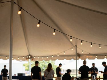 String lights being hung along tent perimeter poles by a crew member, late afternoon natural light, warm glow beginning to show against canvas ceiling