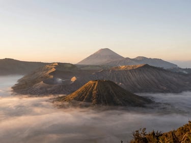 Wide environmental shot of Mount Bromo caldera at sunrise, soft morning mist blanketing the valley, warm golden backlighting, minimalist composition with vast negative space.