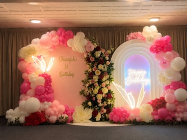 Elegant first birthday backdrop with pink balloon arch, white floral accents, and neon birthday signs.