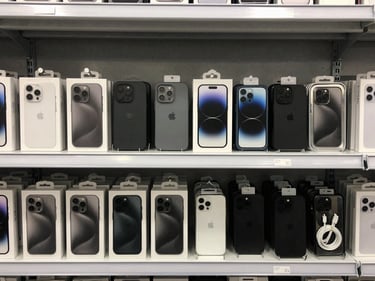 Overhead of assorted mobile accessories — phone cases, screen protectors, charging cables in retail packaging — arranged in neat rows on a light grey warehouse shelf, even lighting, no people