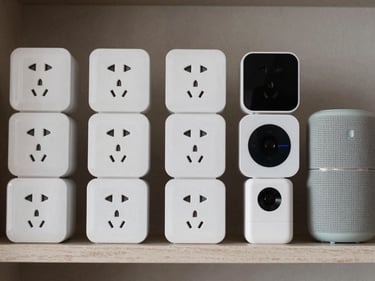 Overhead close-up of smart home devices — smart plugs, mini speakers, tracking devices — arranged in organized grid on a neutral shelf under studio lighting, no people