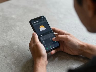 Over-the-shoulder view of hands holding a phone showing a weather data screen, concrete desk surface, flat studio daylight, no people faces